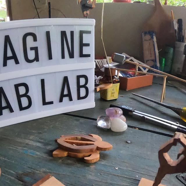 Letreiro luminoso com o nome Imagine Fab Lab sobre a bancada, cercado por ferramentas de entalhe e peças de madeira recortadas.